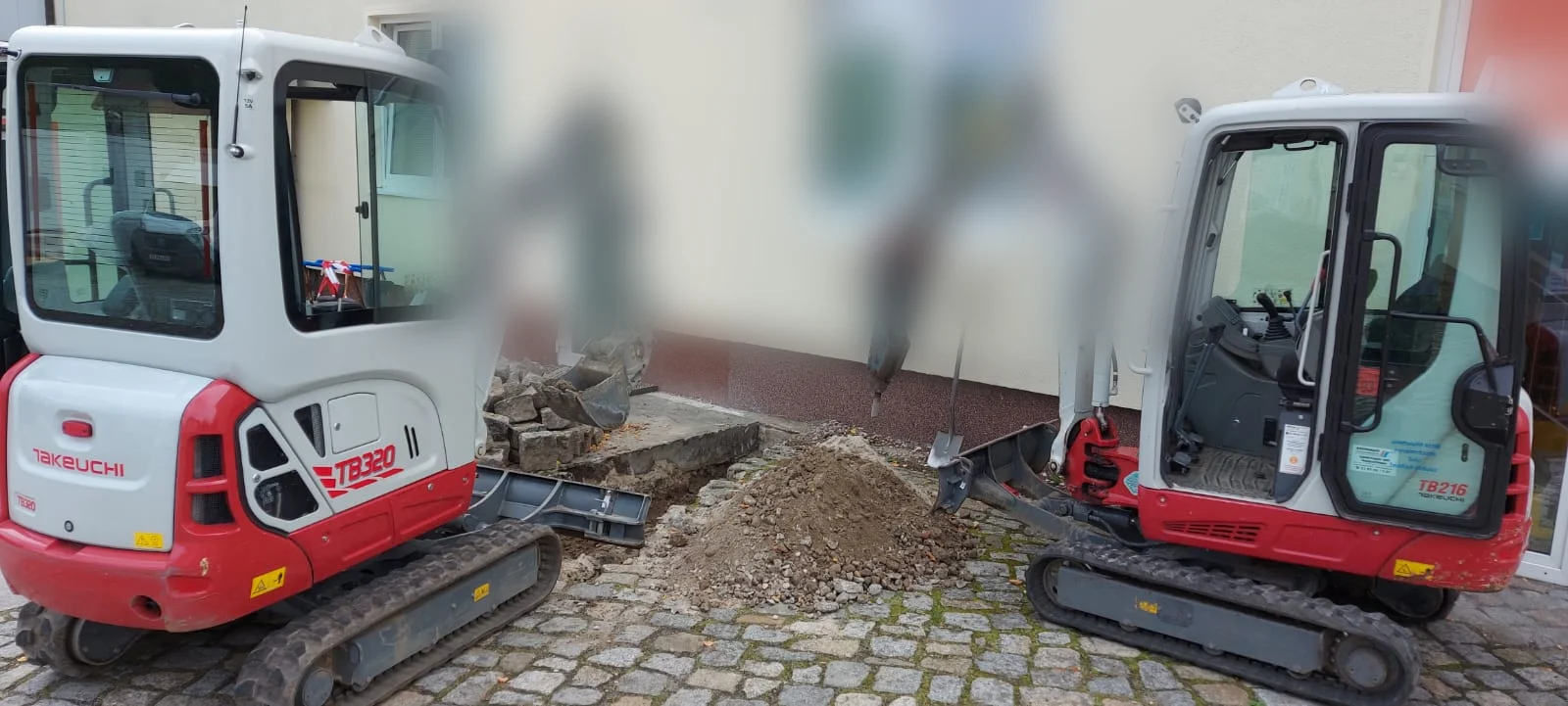 Zwei Minibagger nebeneinander beim Aufbrechen einer Pflasterfläche an einer Hauswand